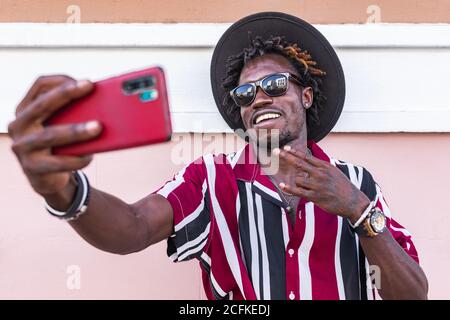 Funky young African American guy in stylish outfit and accessories standing near colorful wall and taking selfie on smartphone on urban street Stock Photo
