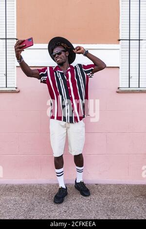 Funky young African American guy in stylish outfit and accessories standing near colorful wall and taking selfie on smartphone on urban street Stock Photo