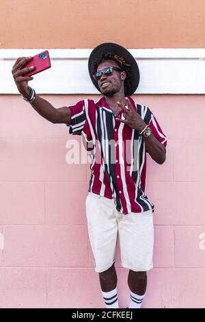 Funky young African American guy in stylish outfit and accessories standing near colorful wall and taking selfie on smartphone on urban street Stock Photo