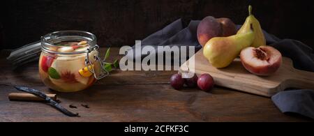 Glass jar with canned fruits and fresh pears, peaches and plums on a dark rustic wooden table, moody vintage still life style in panoramic format, cop Stock Photo