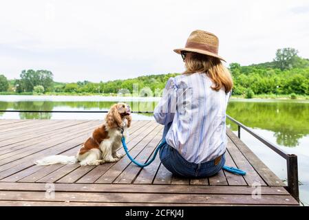 Rear view shot of woman wearing straw hat and casual clothes while sitting on the pier next to her puppy and looking each other. Copy space. Stock Photo
