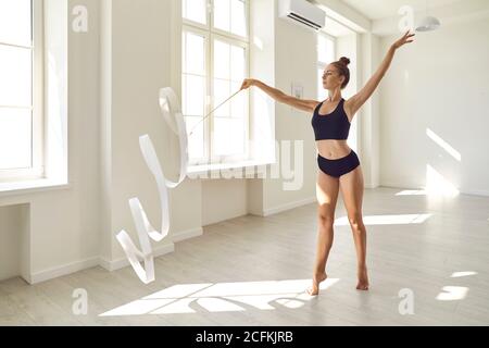 Beautiful young gymnast with white ribbon rehearsing routine moves in modern dance studio Stock Photo