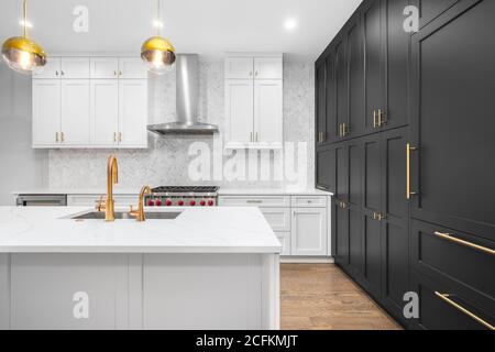 A luxurious, modern kitchen with white and black cabinets, gold hardware/faucets, and white herringbone marble tiles. Stock Photo