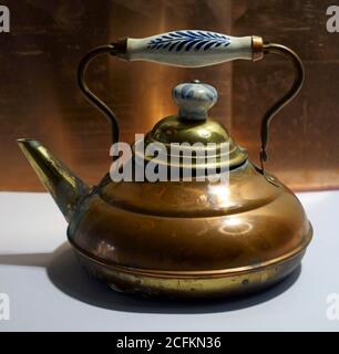 Old often repaired copper kettle. The blue white grip indicates a Dutch origin. In the background is a copper plate. Stock Photo