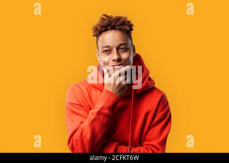 Freestyle. Mulatto guy in hoodie standing isolated on yellow touching chin smiling playful Stock Photo