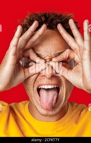 Freestyle. Mulatto freckled guy in yellow t-shirt standing isolated on dark pink making eyeglasses with hands showing tongue playful face close-up Stock Photo