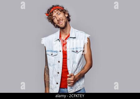 Freestyle. Man with curly hair in jean sleeveless jacket and headband standing isolated on grey posing smiling joyful Stock Photo