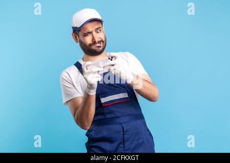 Funny surprised handyman in overalls and gloves holding mobile phone, reading shocking message, crazy online order to deliver something unbelievable, Stock Photo