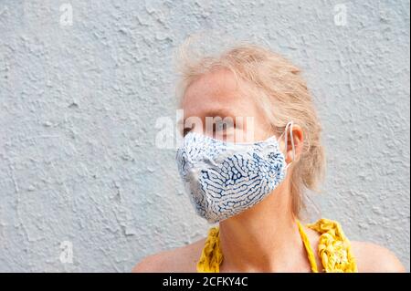 Calm middle aged female wearing protective mask standing on street near concrete building and looking away during coronavirus epidemic Stock Photo