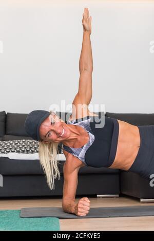 Strong female athlete in sportswear doing side plank exercise while smiling and training at home Stock Photo