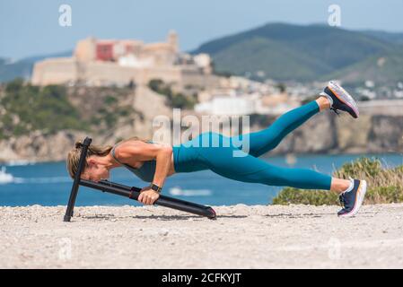 Side view of strong female athlete doing push ups on metal equipment on seashore in summer during intense training Stock Photo