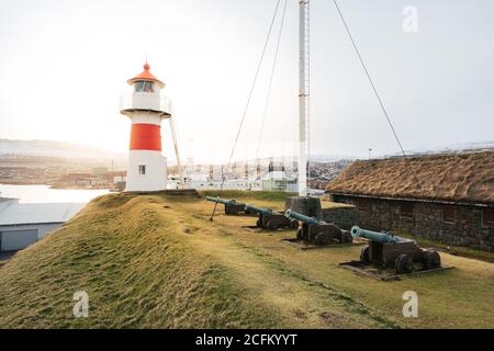 Picturesque view of shabby cannons placed in row on grassy hill with lighthouse on background of gray sky on Faroe Islands in autumn Stock Photo