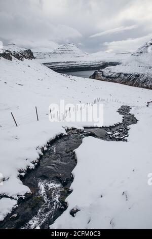 Narrow creek with cold water flowing under snow against mountains and overcast sky on Faroe Island Stock Photo