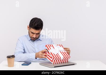 Curious man employee sitting in office workplace, opening gift box and looking inside, unwrapping present, bonus for successful job project, celebrati Stock Photo