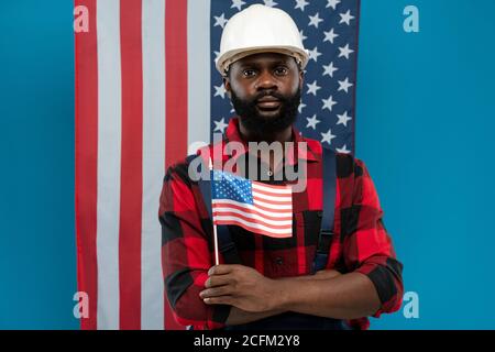 Young bearded mechanic or repairman in flannel and overalls holding small flag Stock Photo