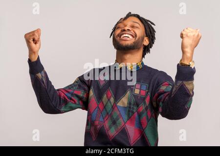 Hurray! Extremely satisfied happy african man with dreadlocks rising hands up with toothy smile on face, pleased, amazed with victory, success. Indoor Stock Photo