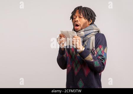 Upset african man with dreadlocks in checkered scarf widely open mouth going to sneeze, seasonal allergy, flu. Indoor studio shot isolated on gray bac