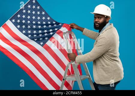 Young bearded African-american worker standing on stepladder and holding US flag Stock Photo