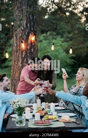 Young joyful friends of various ethnicities sitting by served festive table Stock Photo