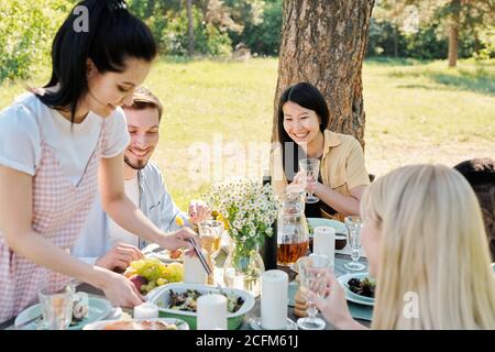 Young cheerful multicultural friends looking at girl mixing cooked vegetables Stock Photo