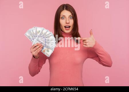 Surprised amazed woman with brown hair pointing finger at fan of dollars in her hand with big eyes and open mouth, shocked with benefits. Indoor studi Stock Photo