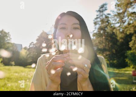 Young Asian woman with long dark hair blowing at dandelion while having fun