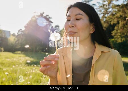 Happy young Asian woman in casualwear blowing at dandelion in her hand Stock Photo