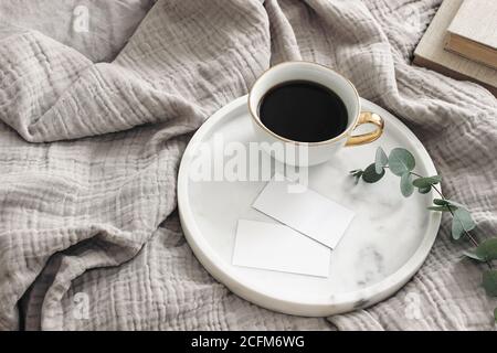 Breakfast in bed stationery mockup scene. Cup of coffee, blank business cards on white marble tray. Green eucalyptus tree branch and books on muslin Stock Photo