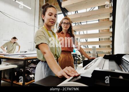 Two young tailors standing by large industrial printer under staircase in studio Stock Photo