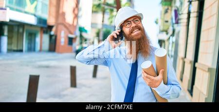 Young redhead architect man with long beard wearing hardhat smiling happy. Having conversation talking on the smartphone holding blueprints at street Stock Photo