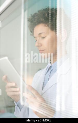 Young female researcher in whitecoat looking through data on tablet screen Stock Photo