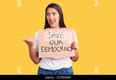 Young beautiful woman holding save our democracy cardboard banner pointing thumb up to the side smiling happy with open mouth Stock Photo