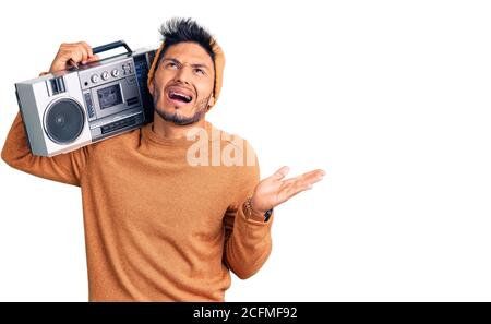 Handsome latin american young man holding boombox, listening to music crazy and mad shouting and yelling with aggressive expression and arms raised. f Stock Photo