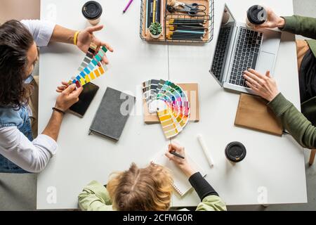 Overview of three young creative designers sitting by desk and choosing colors Stock Photo
