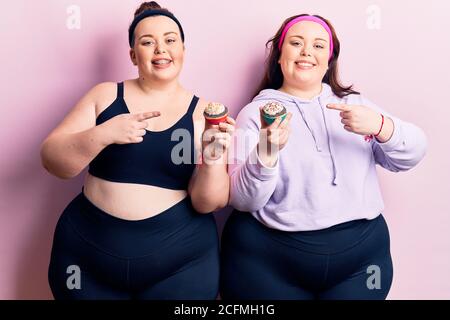 Young plus size twins wearing sportswear holding cupcake smiling happy pointing with hand and finger Stock Photo