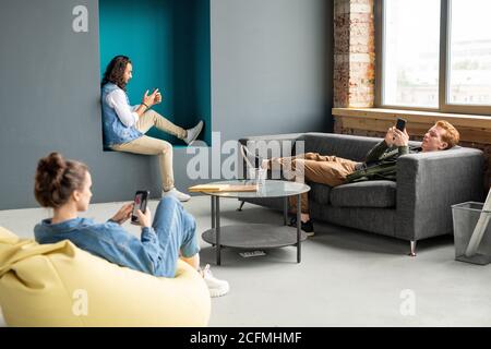 Three young restful specialists using mobile gadgets while relaxing in office Stock Photo