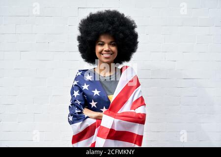 Happy African woman stand on white background wrapped in USA flag, portrait. Stock Photo