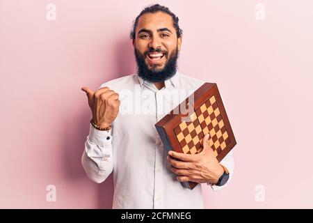 Young arab man holding chess pointing thumb up to the side smiling happy with open mouth Stock Photo