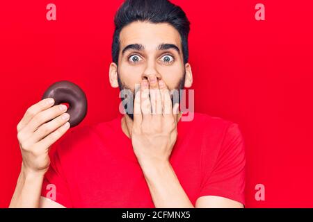 Young handsome man with beard holding donut covering mouth with hand, shocked and afraid for mistake. surprised expression Stock Photo