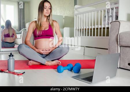 portrait of young beautiful caucasian pregnant woman sitting on yoga mat watching workout tutorial at home, copy space. Healthy pregnancy. Stock Photo