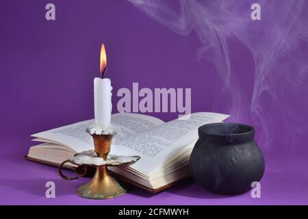 Candle burning in candlestick and witch smoky pot and on the dark purple smoked background with an opened book. Halloween concept Stock Photo