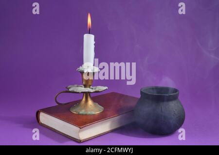 Candle in candlestick burning on an old book and witch smoky pot on the dark purple smoked background. Halloween concept Stock Photo