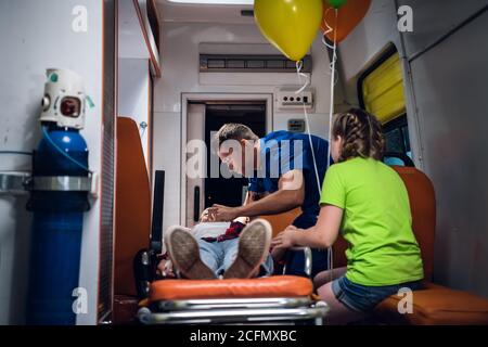 Unconscious mother lying on a stretcher in an ambulance car, a paramedic providing her first aid. Stock Photo