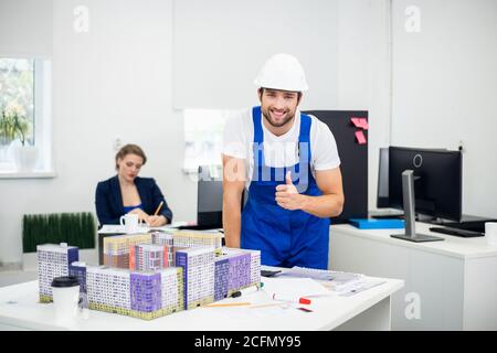 Young male architect working in the office. He is leaning on the table with a large construction maquette on it and showing thumbs up. Stock Photo