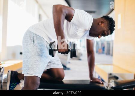 Side view of African American man in sportswear leaning on bench and doing row exercise with dumbbell during training in gym. Stock Photo