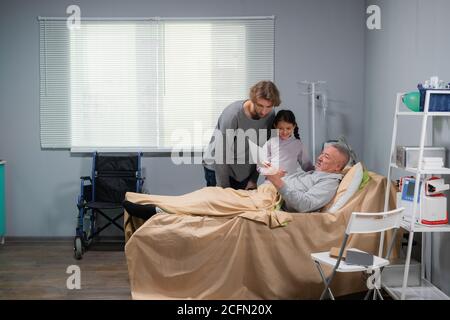 A little girl and her dad are visiting an old man in a nursing home, the man is staying in bed. Stock Photo