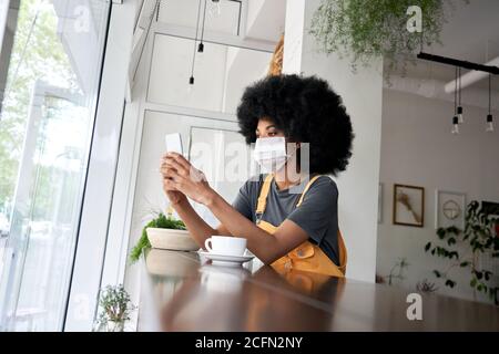 African lady wearing face mask using phone sit at table in modern cafe. Stock Photo