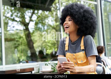 Cheerful African woman using phone, laughing, sitting at outdoor cafe table. Stock Photo