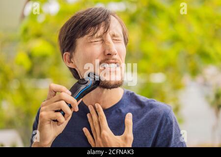 man is shaving off his beard with electric razor on the balcony during quarantine. Handsome bearded man trimming his beard with a trimmer at home Stock Photo