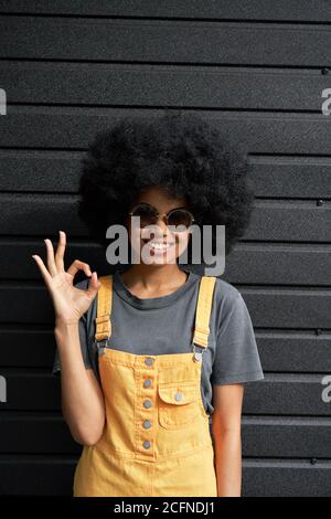Happy African hipster lady wear stylish glasses stand against black wall. Stock Photo